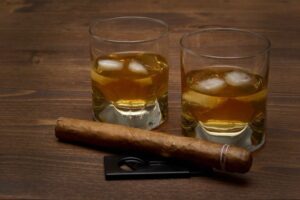 two glasses of whiskey with ice cubes on table with cigar representing long distance move of alcohol tobacco and firearms
