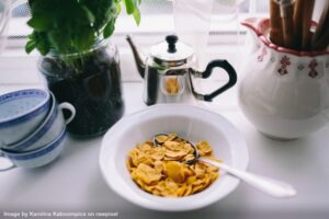 a bowl of cereal on a table beside a pitcher, a plant, a stainless steel coffee pot and some coffee cups representing a morning routine to boost productivity